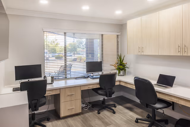 A small office space with three black swivel chairs positioned at a light wood L-shaped desk. Two computer monitors and a laptop are on the desk, along with a telephone and a potted plant. A large window with horizontal blinds looks out onto an outdoor area with trees and parked cars. The room has light-colored walls and wood flooring.