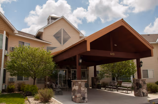 Entrance of a senior living facility with a covered driveway supported by stone pillars, benches, chairs, and trees on either side under a partly cloudy sky.