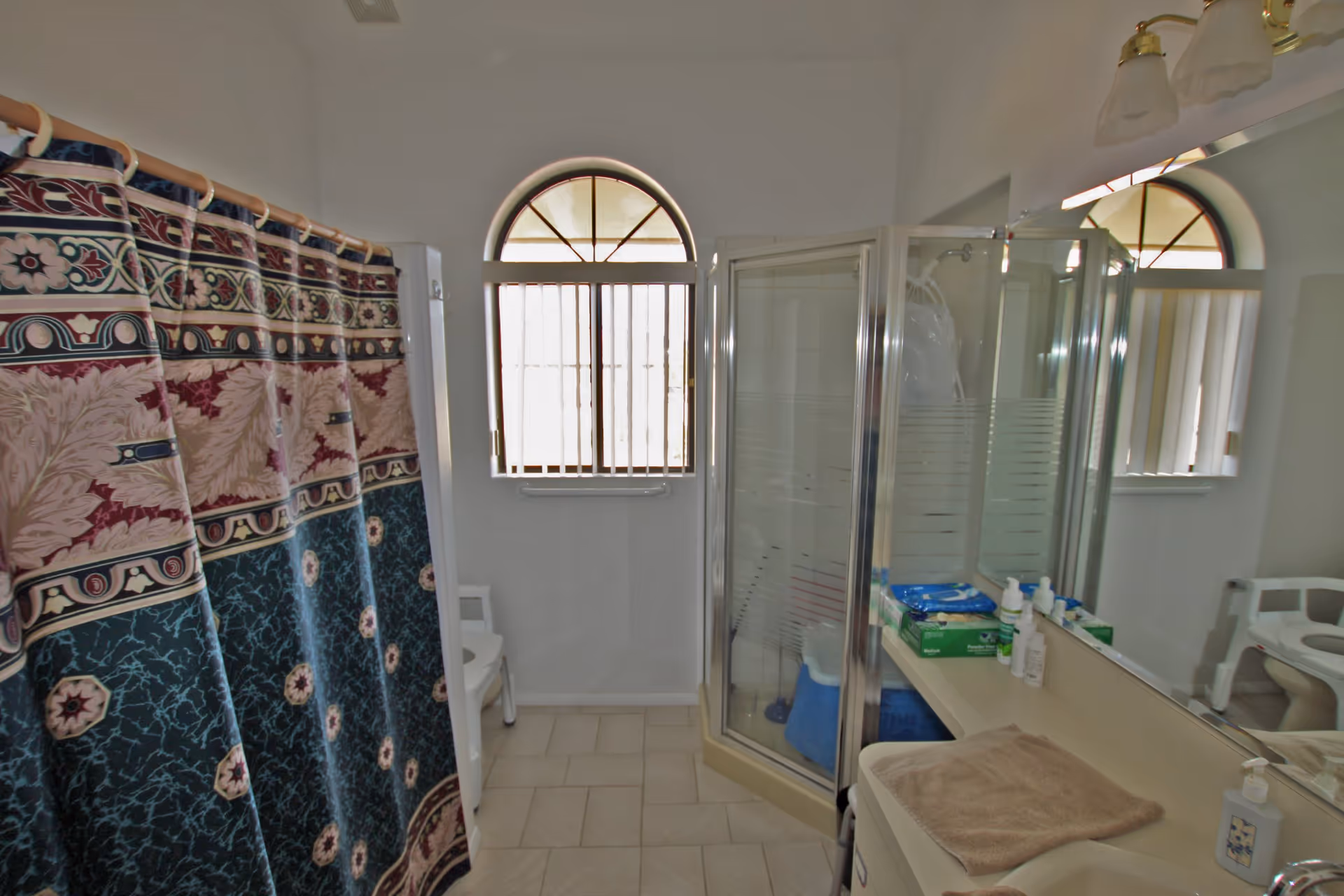 Bathroom with a patterned shower curtain on the left, a glass-enclosed shower stall in the center, a window with an arched top and vertical blinds, a white chair near the window, and a countertop with toiletries and a towel on the right.