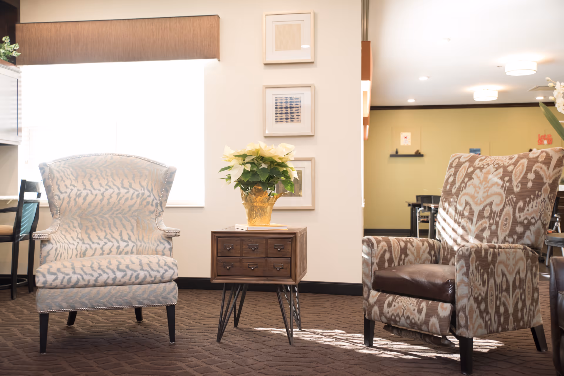 A cozy sitting area with two patterned armchairs separated by a small wooden side table holding a potted plant with white flowers. The room has a carpeted floor, light-colored walls with three framed pictures, and soft natural light coming through a window with a brown valance.