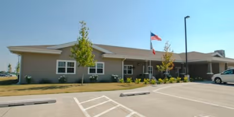 Single-story beige senior living facility building with an American flag at the entrance and a parking lot in front.