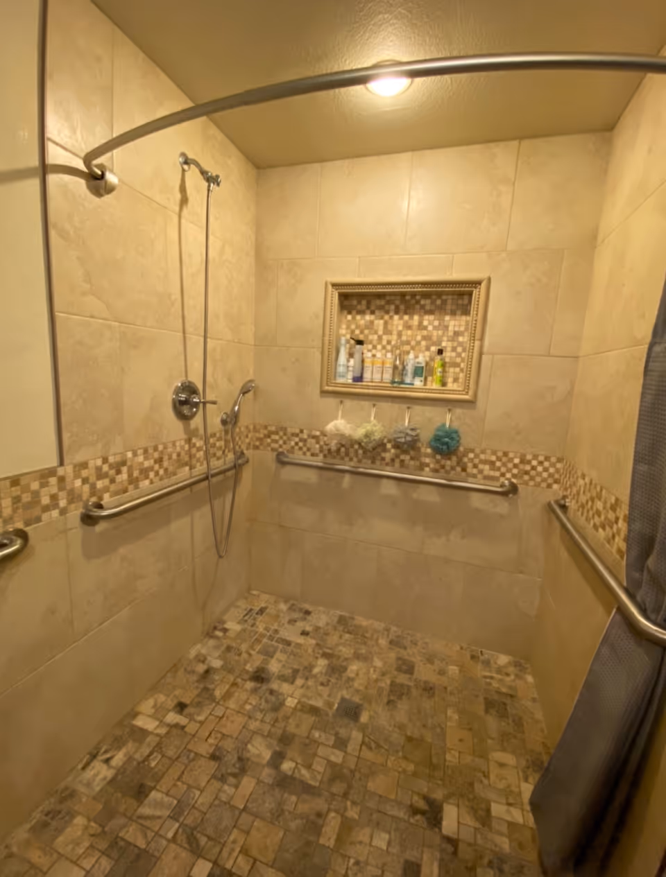 Tiled walk-in shower with grab bars, a handheld showerhead, a recessed shelf of toiletries and hanging bath poufs.