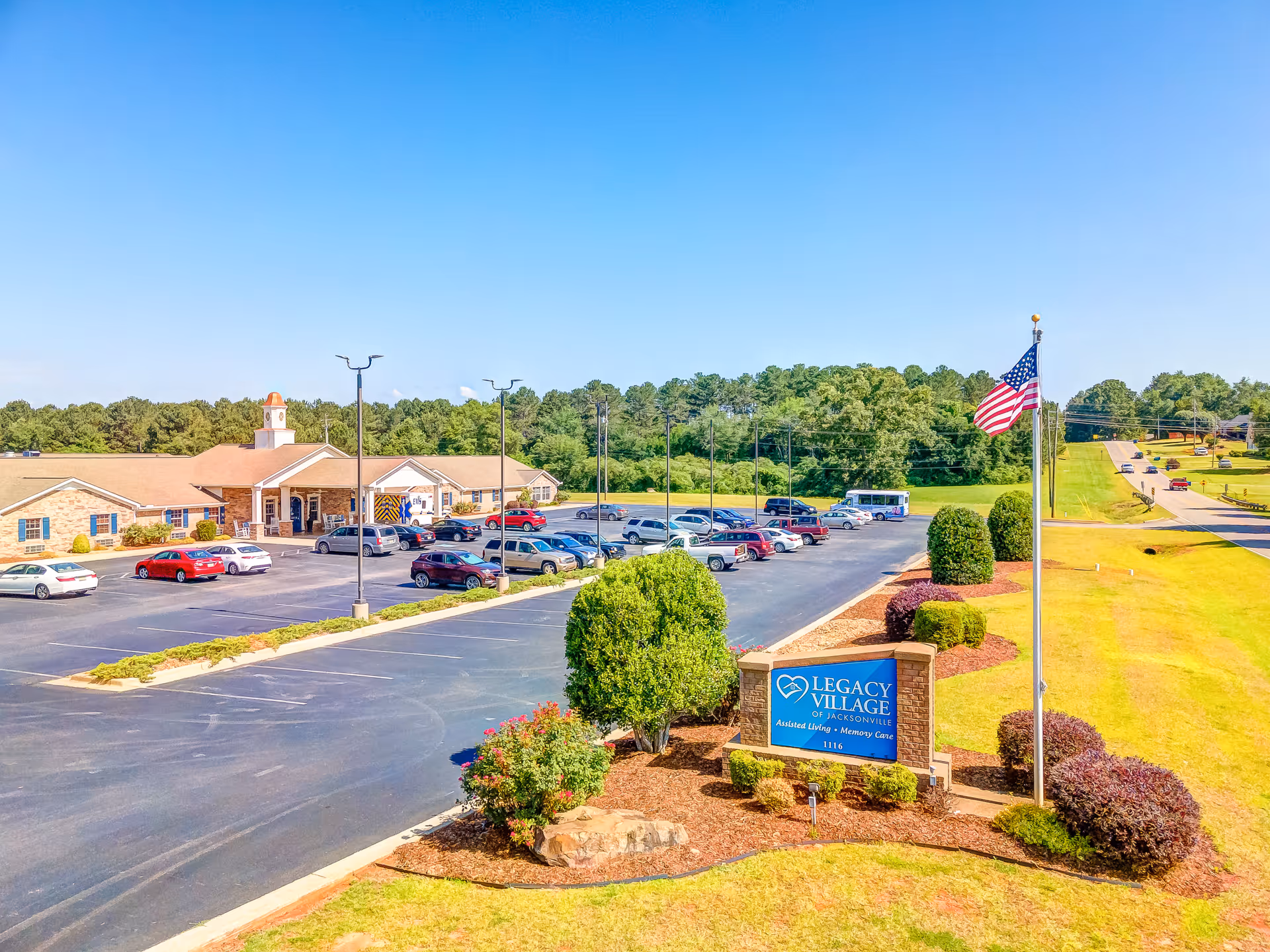 Exterior view of Legacy Village of Jacksonville senior living facility with a parking lot filled with cars, a flagpole with an American flag, and a sign displaying the facility's name. The building is surrounded by green trees and landscaping under a clear blue sky.