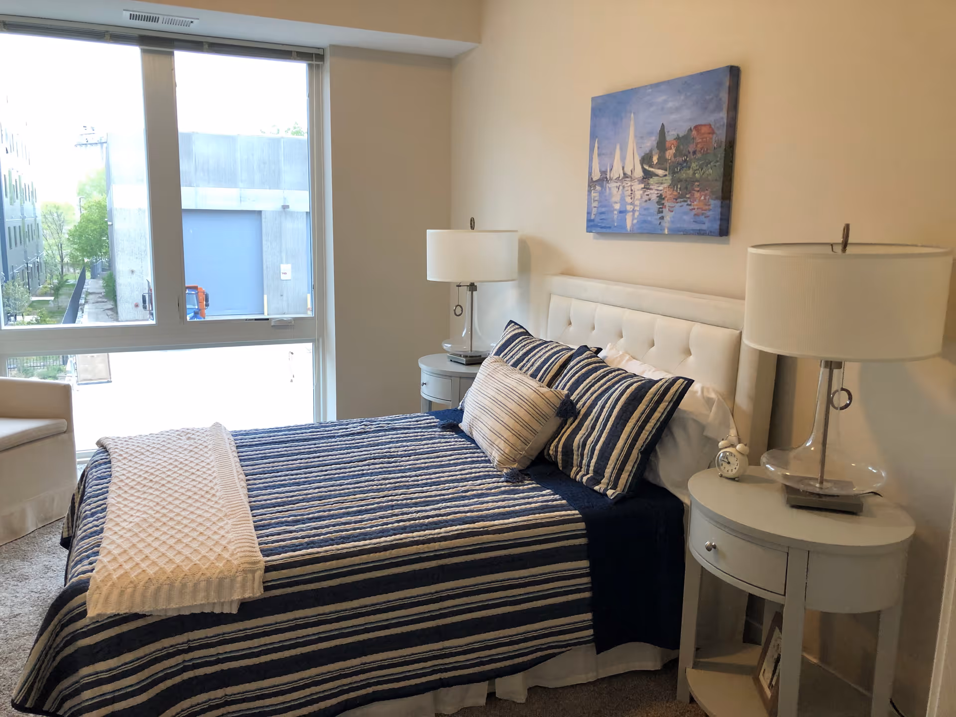 A bedroom with a bed covered in a blue and white striped comforter and matching pillows. There are two round bedside tables, each with a lamp and one with a small clock. A painting of sailboats on water hangs above the bed. A large window lets in natural light, and a beige armchair is partially visible to the left.