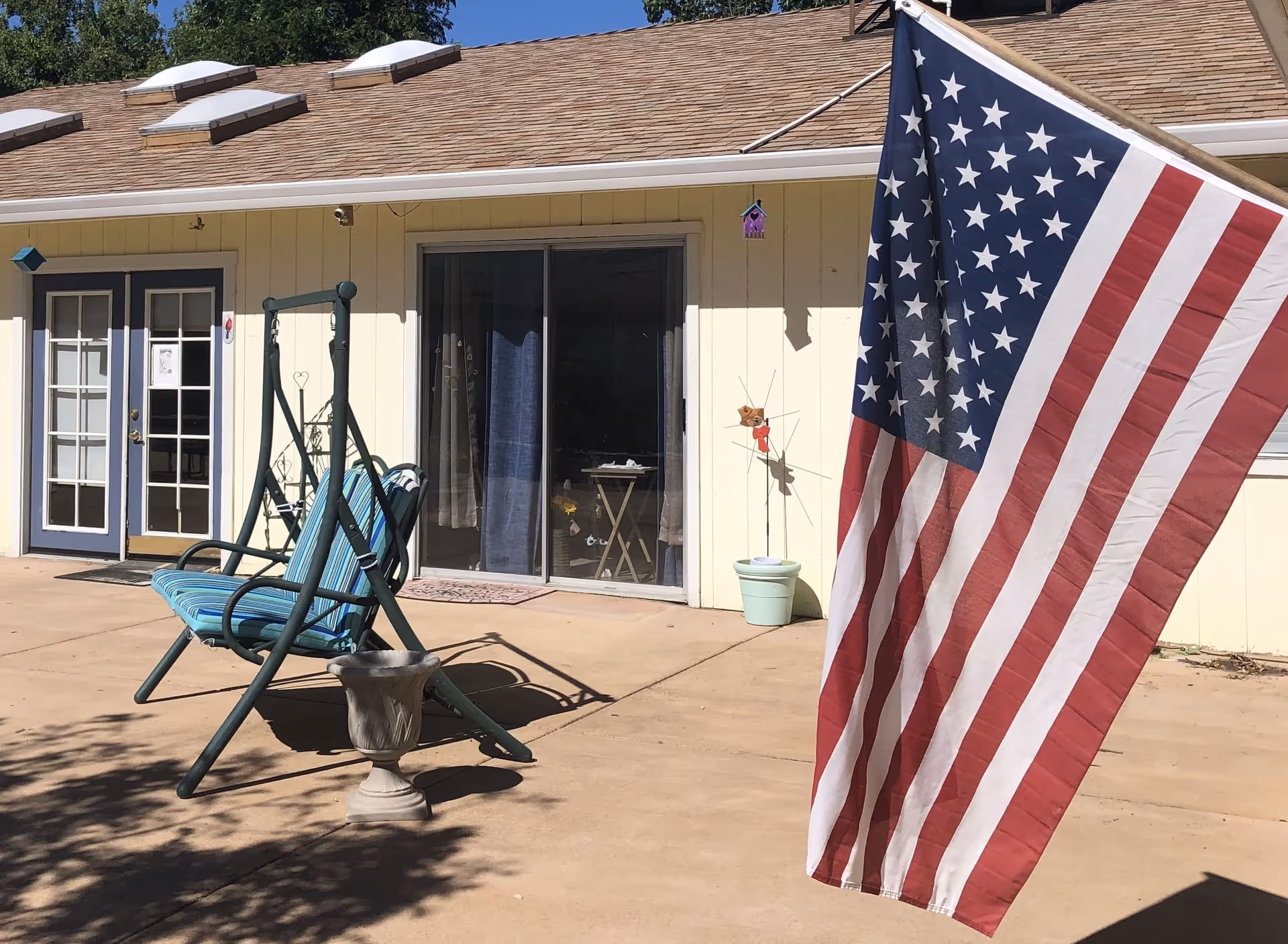 Outdoor patio area with a green metal swing bench with blue striped cushions, a large American flag hanging on the right side, a beige building with sliding glass doors and a double door with glass panes in the background, and a small potted plant on the ground.