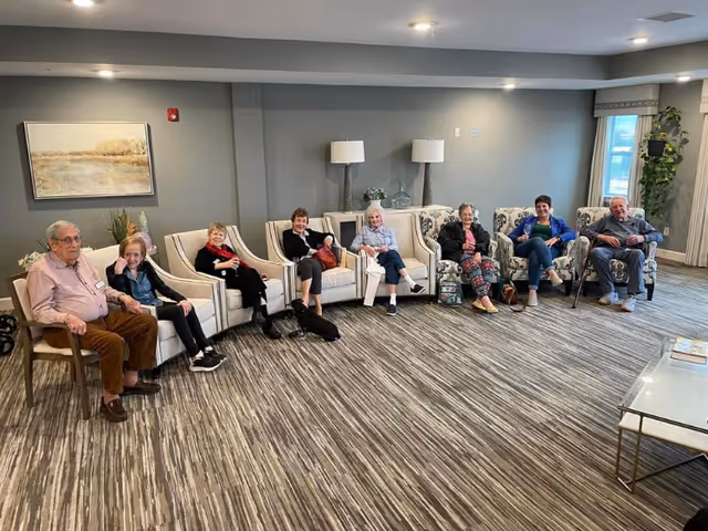 A group of eight elderly people sitting in a semi-circle on armchairs in a well-lit room with gray walls and carpeted floor. There are two lamps and decorative items on a table against the wall, a large window with curtains, and a framed landscape painting on the wall. Some of the individuals have walking aids nearby.