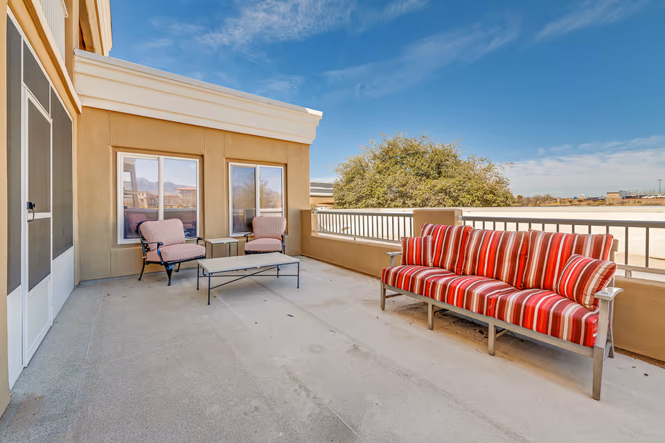 Sunlit outdoor terrace with a red-striped sofa, two cushioned chairs, and a low coffee table beside a building.