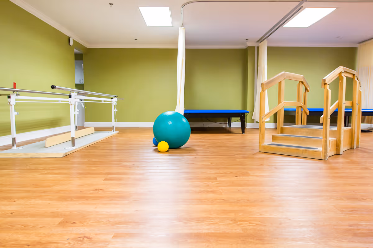 A physical therapy room with wooden flooring, green walls, parallel bars for walking exercises on the left, a set of wooden stairs with handrails on the right, and exercise balls in the center. The room is well-lit with ceiling lights and has a blue padded bench against the back wall.