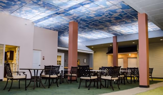 Indoor seating area with multiple black metal tables and chairs with beige cushions, supported by reddish-brown pillars. The ceiling features a sky and clouds design, creating an outdoor illusion. Surrounding walls resemble house exteriors with doors and windows.