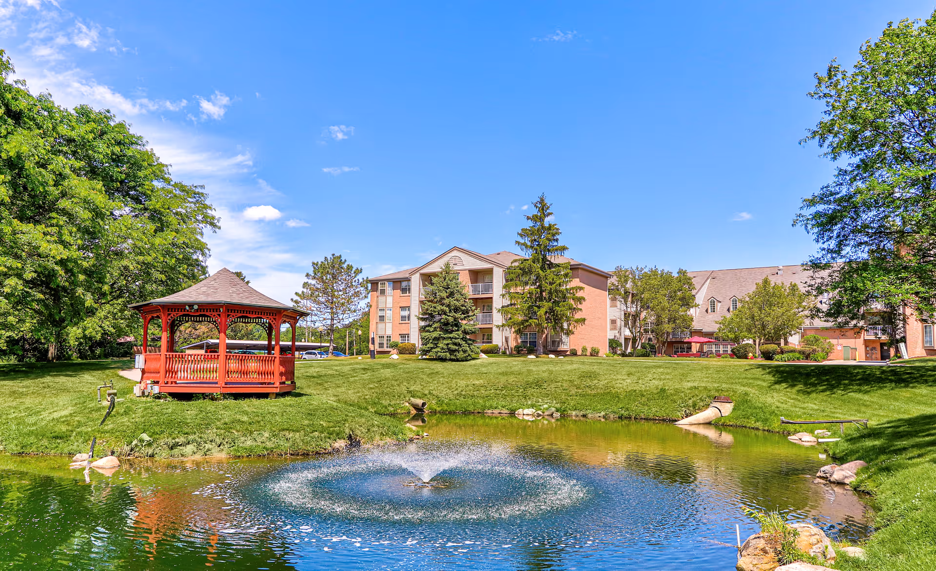A scenic outdoor area at Rosehaven Manor featuring a small pond with a water fountain, a red wooden gazebo on the grassy bank, surrounded by green trees and a multi-story residential building in the background under a clear blue sky.