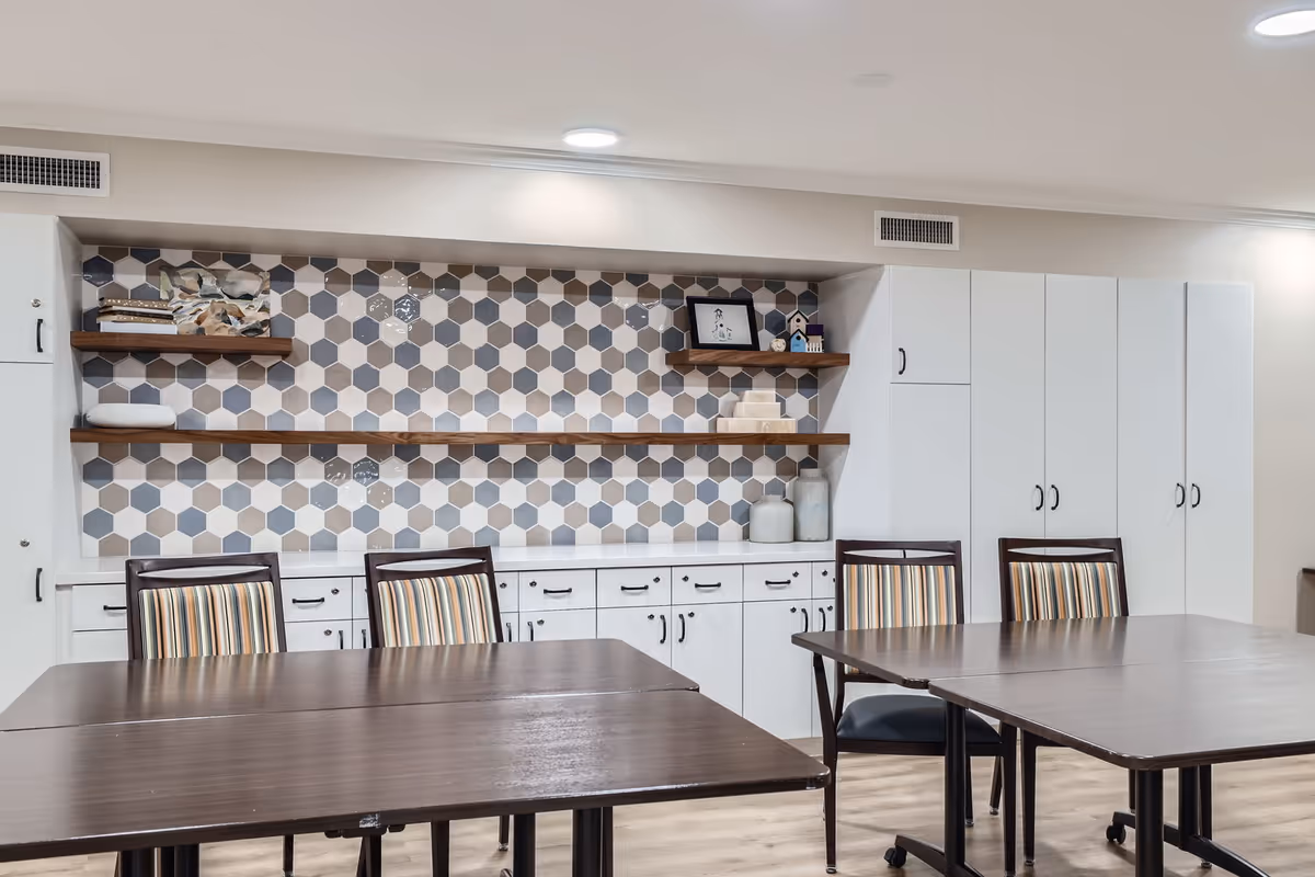 Interior view of a room with two dark wooden tables and four chairs with striped upholstery. The back wall features white cabinets and drawers with black handles, two wooden floating shelves displaying decorative items, and a hexagonal patterned tile backsplash in shades of gray, beige, and white. The floor is light wood, and the ceiling has recessed lighting and air vents.