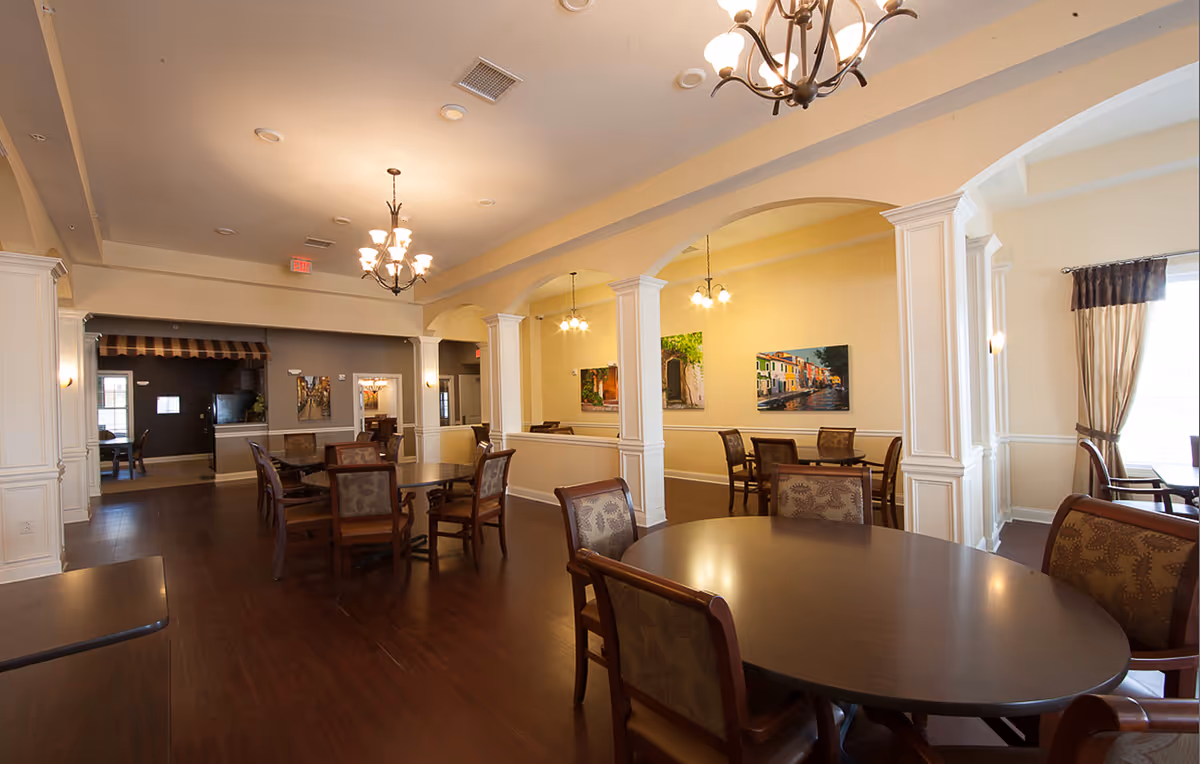 Spacious dining room in a senior living facility with multiple wooden tables and chairs arranged neatly. The room features warm lighting from chandeliers and pendant lights, cream-colored walls with decorative columns and arches, and framed artwork on the walls. Large windows with curtains allow natural light to enter the space.