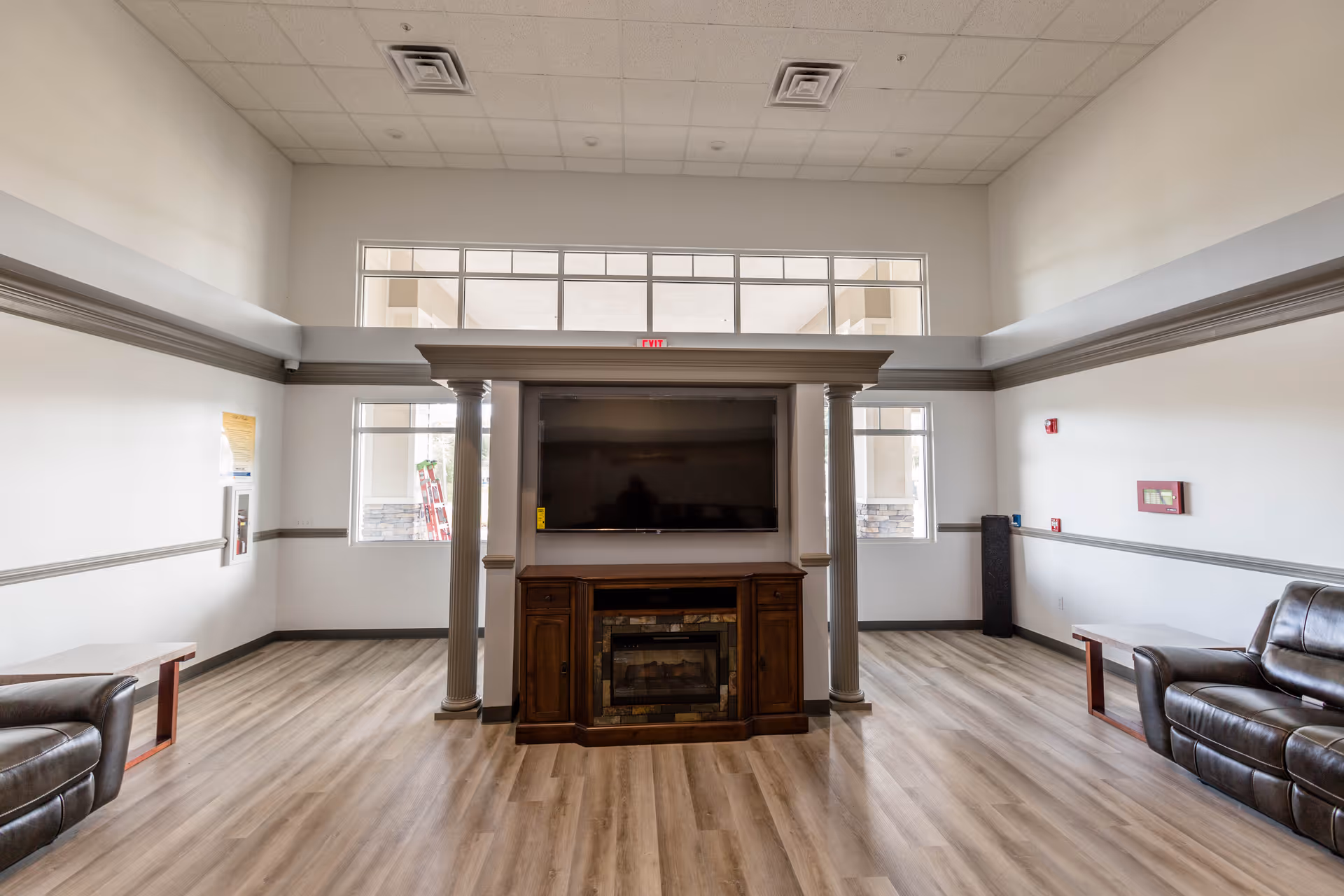 A spacious living room area with light wood flooring, white walls, and a high ceiling with recessed lighting. The room features a large flat-screen TV mounted above a wooden electric fireplace, flanked by two columns. There are leather sofas on either side of the room with small wooden side tables next to them. Large windows near the ceiling and on the walls allow natural light to enter the space.