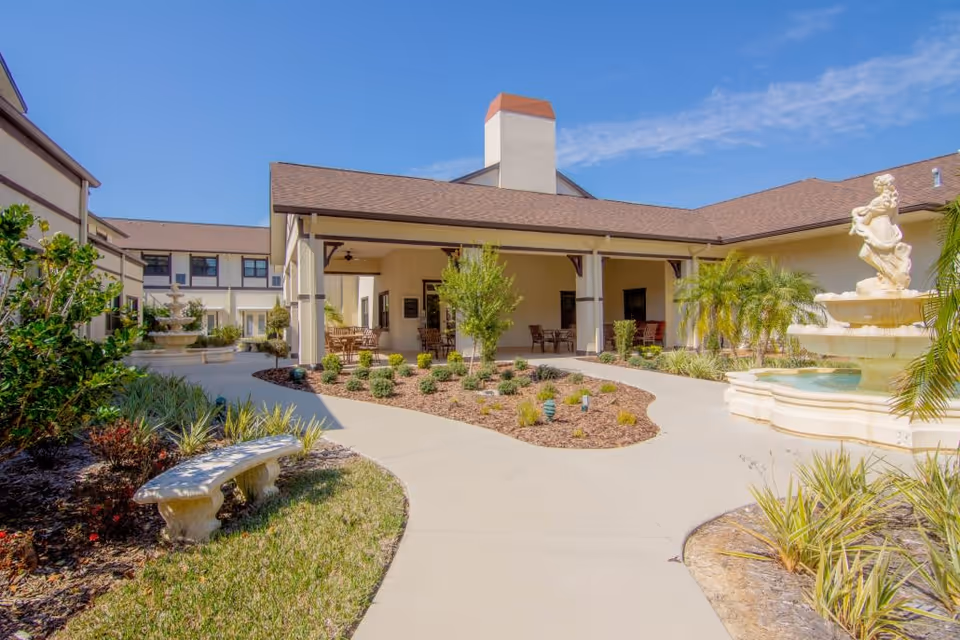Outdoor courtyard area of Royal Dalton House featuring a paved walkway, landscaped garden beds with shrubs and small trees, a stone bench, and a decorative fountain with a statue. The building surrounds the courtyard with covered seating areas and a clear blue sky overhead.