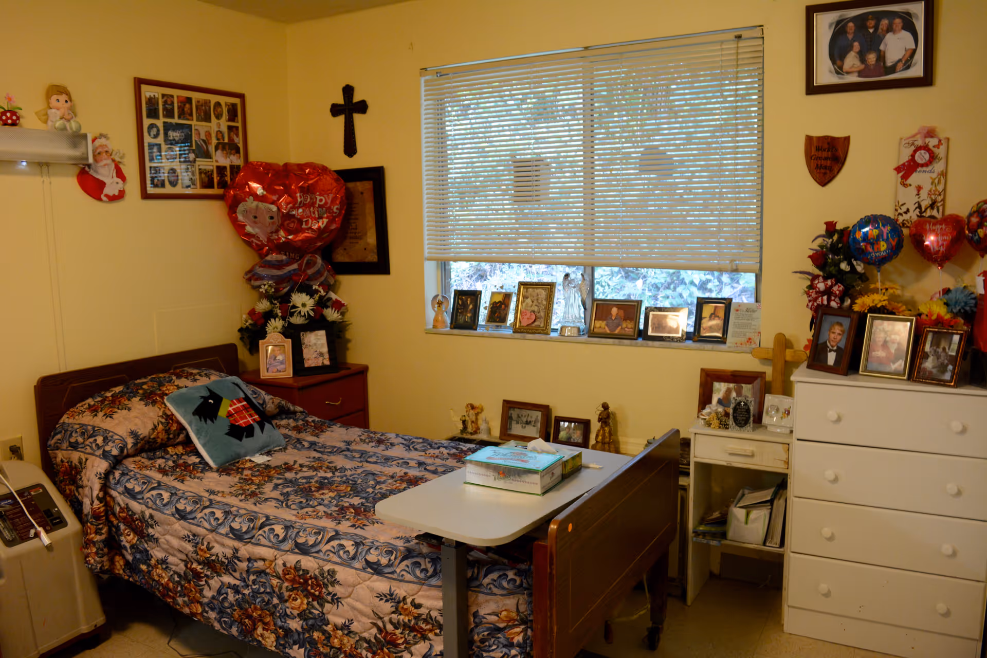 A cozy nursing home bedroom with a single bed covered in a floral patterned bedspread and a pillow with a dog design. There is a bedside table with framed photos and a bouquet of flowers with heart-shaped balloons. A window with blinds is above a shelf filled with framed pictures and small decorative items. A white dresser with more framed photos and balloons is on the right side of the room. The walls are decorated with a cross, plaques, and family photos.