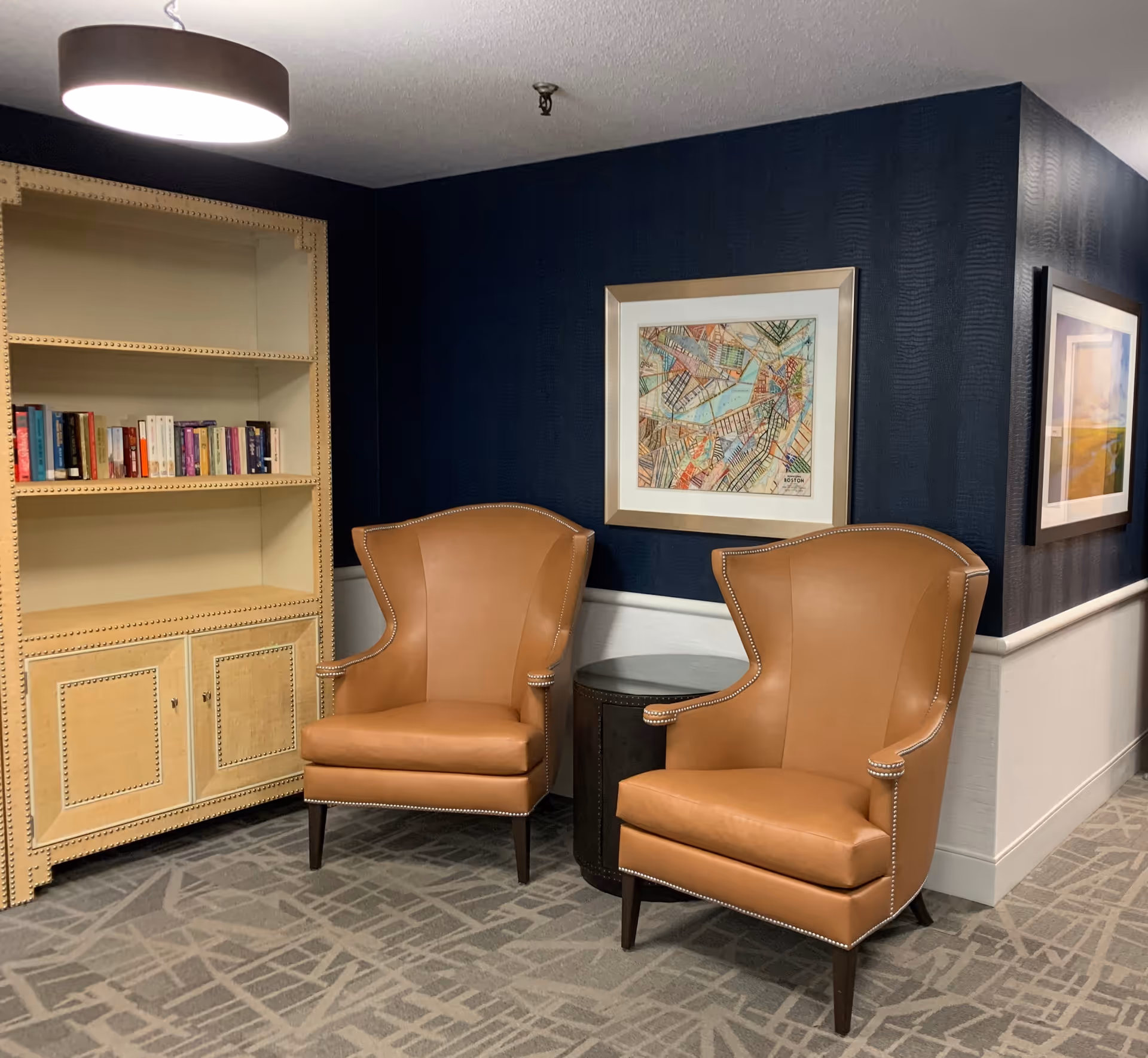 A cozy seating area in a senior living facility with two tan leather wingback chairs positioned around a small round dark side table. Behind the chairs is a dark blue textured wall with two framed artworks hanging. To the left, there is a light-colored wooden bookshelf with a few books on the middle shelf. The floor is carpeted with a geometric pattern, and a modern circular ceiling light hangs above.