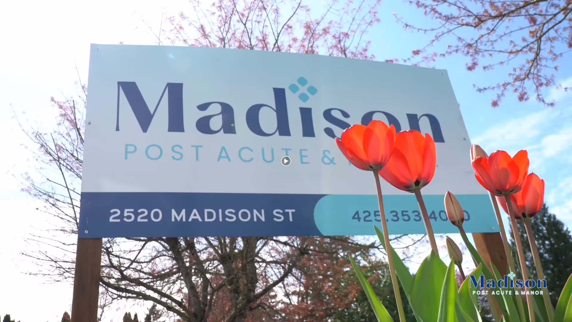 Outdoor sign for Madison Post Acute & Manor located at 2520 Madison St, with red tulips in the foreground and trees in the background under a blue sky.