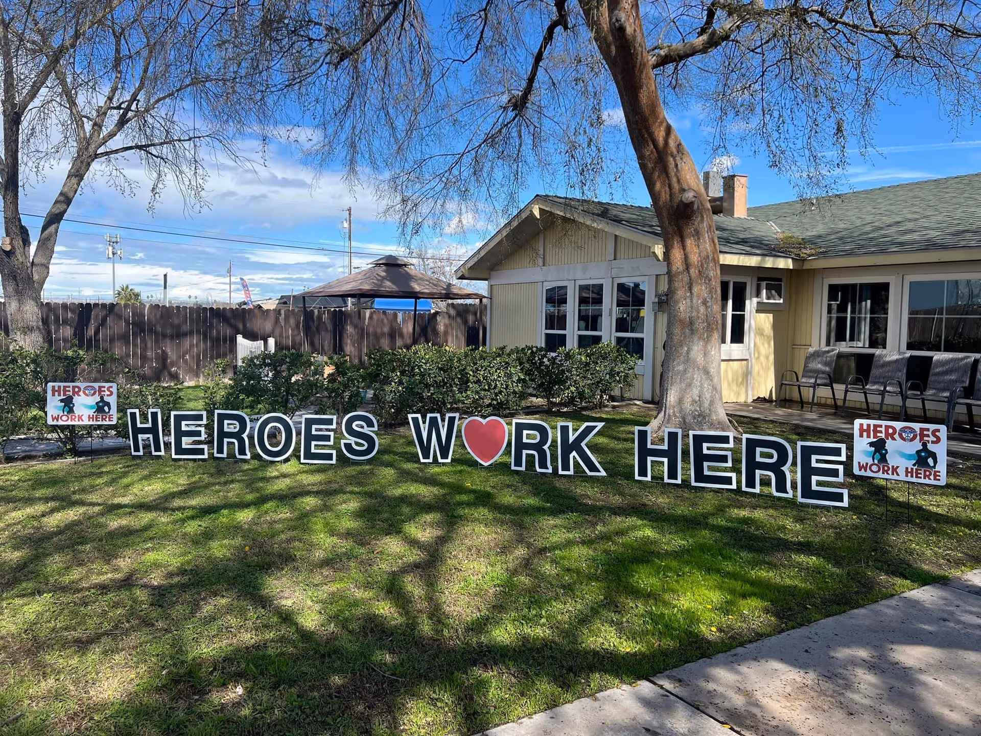 Front lawn of a single-story assisted living building with a large 'HEROES WORK HERE' yard sign and chairs on the porch.