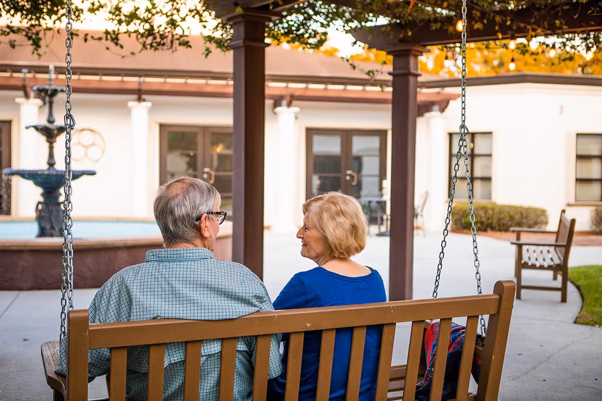 An elderly man and woman sitting on a wooden swing bench in an outdoor courtyard area with a fountain and building in the background. The man is wearing glasses and a green checkered shirt, and the woman is wearing a blue top. They are facing each other and appear to be engaged in conversation.