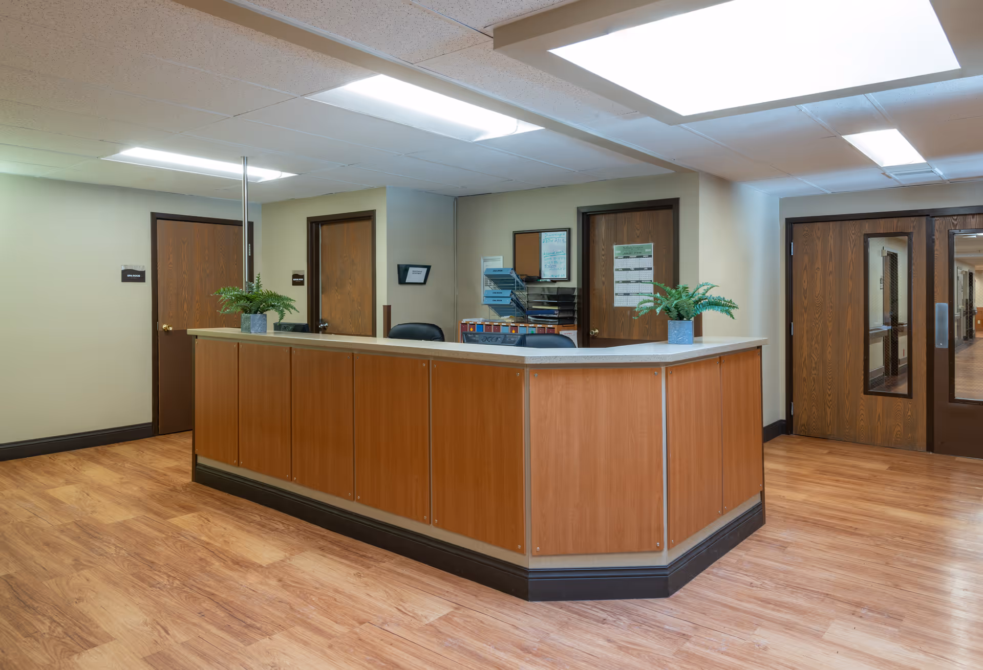 Reception area in Caring Heights Community Care and Rehabilitation Center with a wooden front desk, two potted plants on the counter, wooden doors, and bright overhead lighting.
