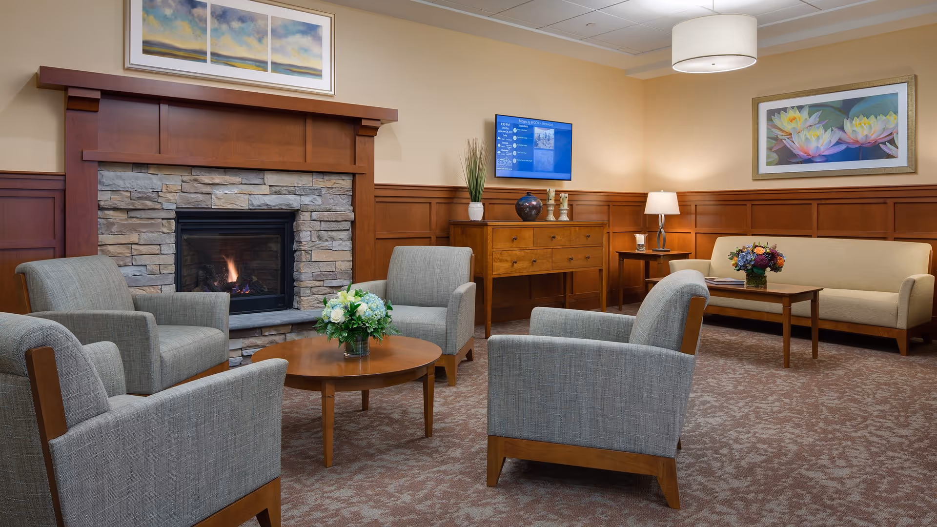 A cozy living room area with a stone fireplace, four gray upholstered armchairs arranged around a round wooden coffee table with a floral arrangement. There is a beige sofa with a wooden coffee table and another floral arrangement, a wooden sideboard with decorative items, a wall-mounted TV, and framed artwork on the walls. The room has warm beige walls and carpeted floor.