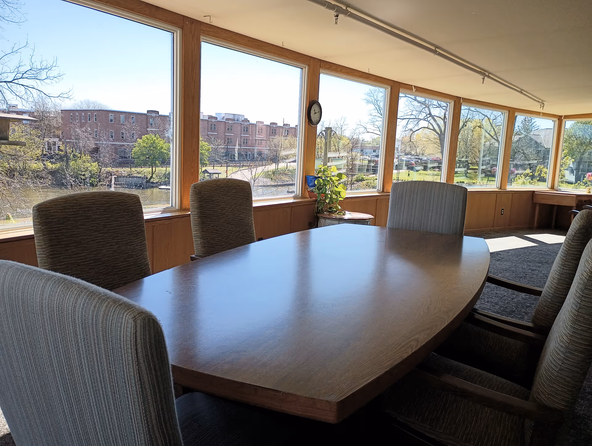 A conference or meeting room with a large wooden table surrounded by six upholstered chairs. The room has large windows along one wall, providing a view of trees, a river, and buildings outside. There is a small round table with a plant and a clock on the wall between the windows.
