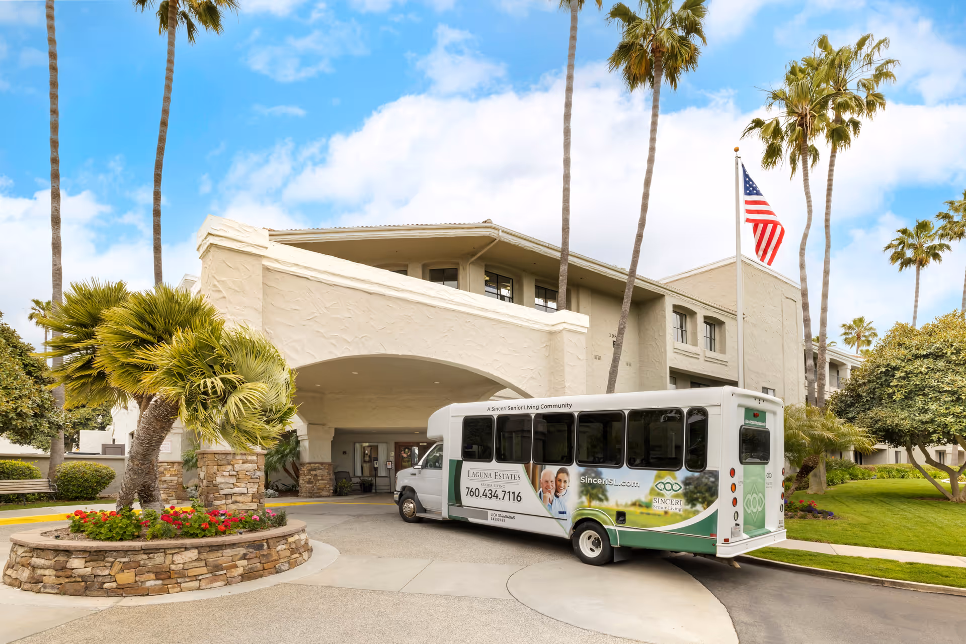 Exterior view of Laguna Estates Senior Living facility with a shuttle bus parked in front. The building has a beige stucco finish with stone accents and is surrounded by palm trees and well-maintained landscaping. An American flag is flying on a flagpole near the entrance.