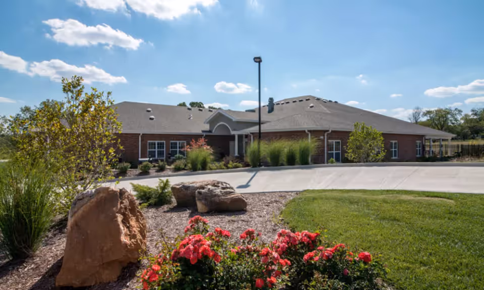 Single-story brick senior living building with a landscaped front entrance, rocks and flowering bushes under a blue sky.
