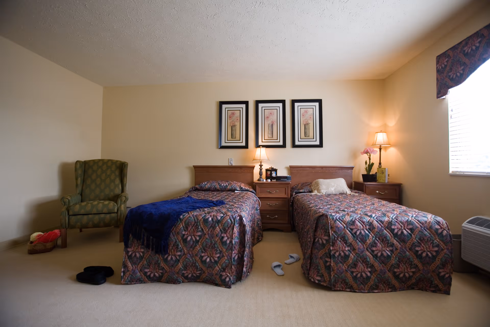 A bedroom with two twin beds covered in floral patterned bedspreads. Between the beds is a wooden nightstand with a lamp, clock, and small decorative items. Above the nightstand are three framed floral pictures. To the left of the beds is a green upholstered armchair with a basket of yarn on the floor nearby. On the floor near the beds are two pairs of slippers. A window with blinds and a valance is on the right wall, and a floor lamp with a flower pot is next to the right bed.