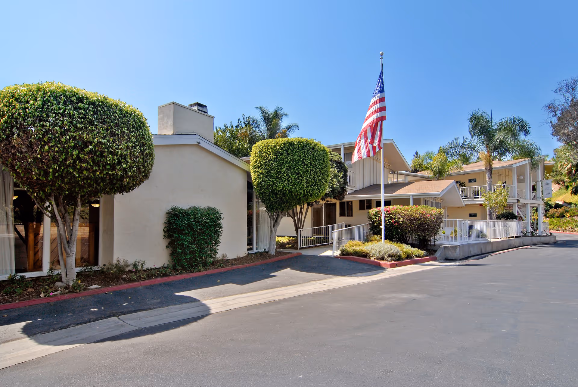 Exterior view of a senior living facility building with neatly trimmed trees, an American flag on a flagpole, and a clear blue sky.