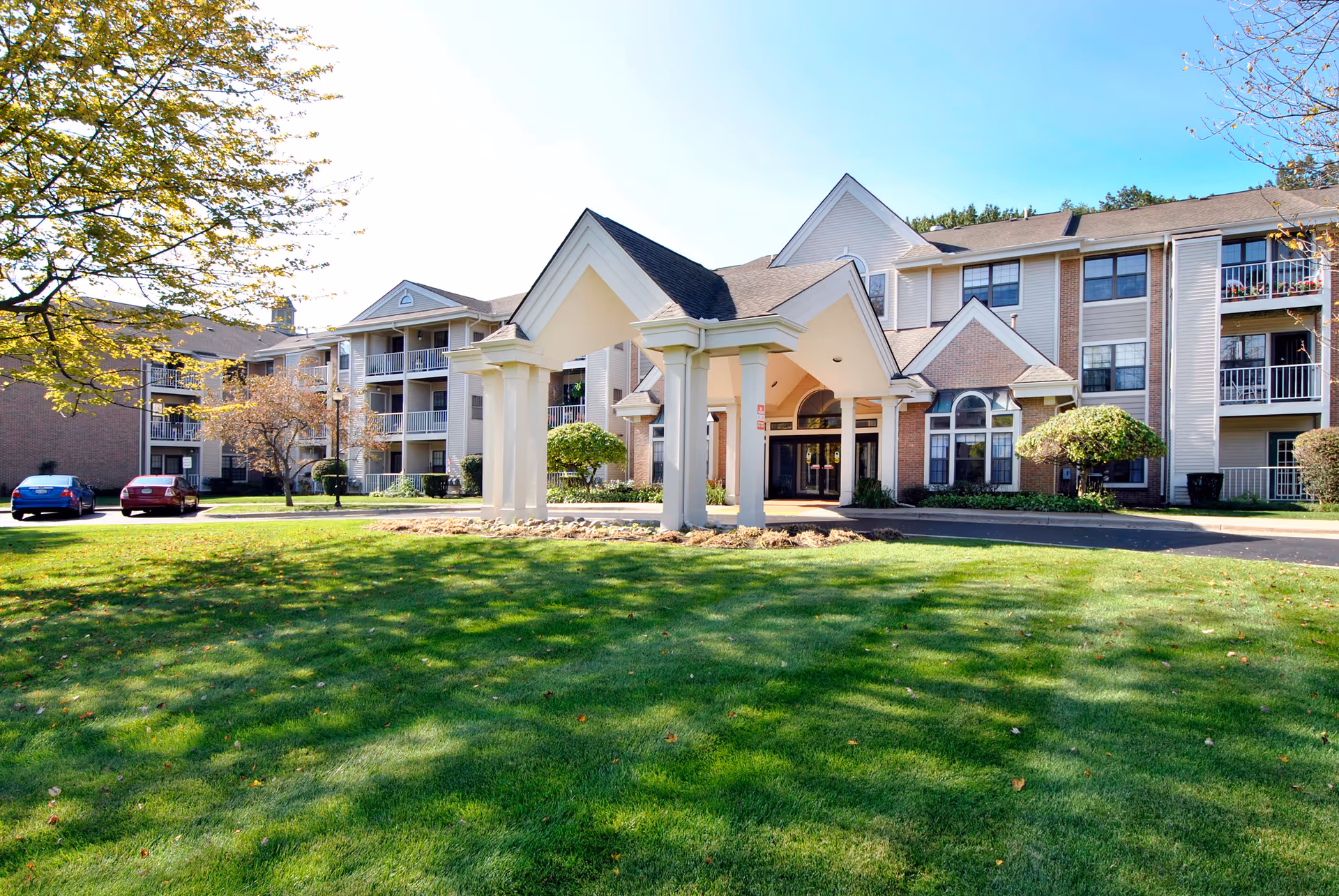 Exterior view of Porthaven Manor, a multi-story senior living facility with a covered entrance, surrounded by green lawn and trees under a clear blue sky.