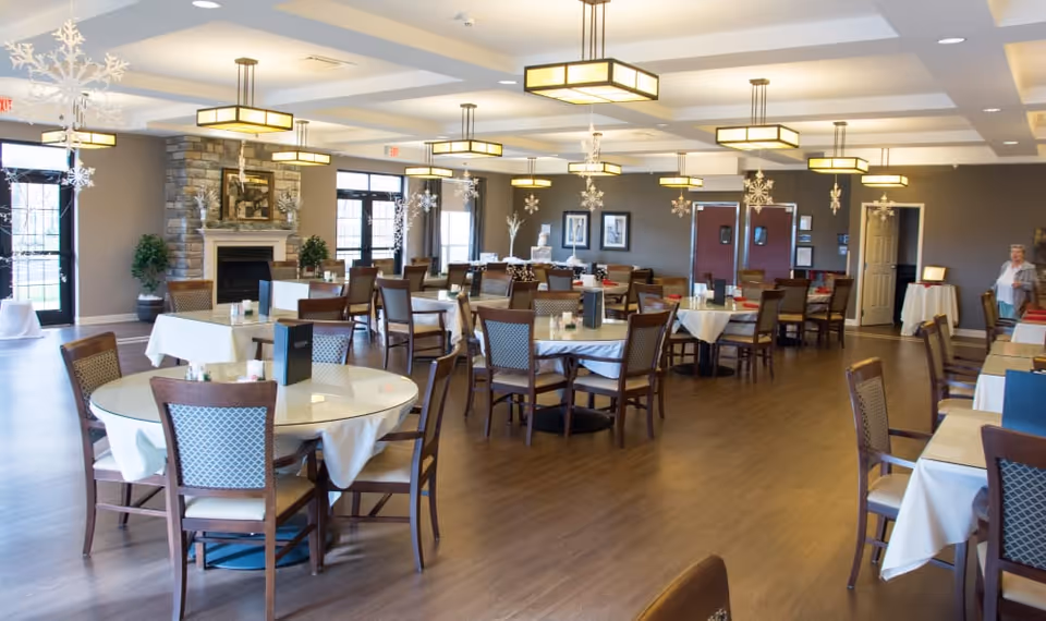 A spacious dining room in a senior living facility with multiple round and square tables covered with white tablecloths, surrounded by wooden chairs with cushioned seats. The room features modern hanging light fixtures, large windows, a stone fireplace, and decorative snowflake ornaments hanging from the ceiling. A person is visible in the background near the entrance.