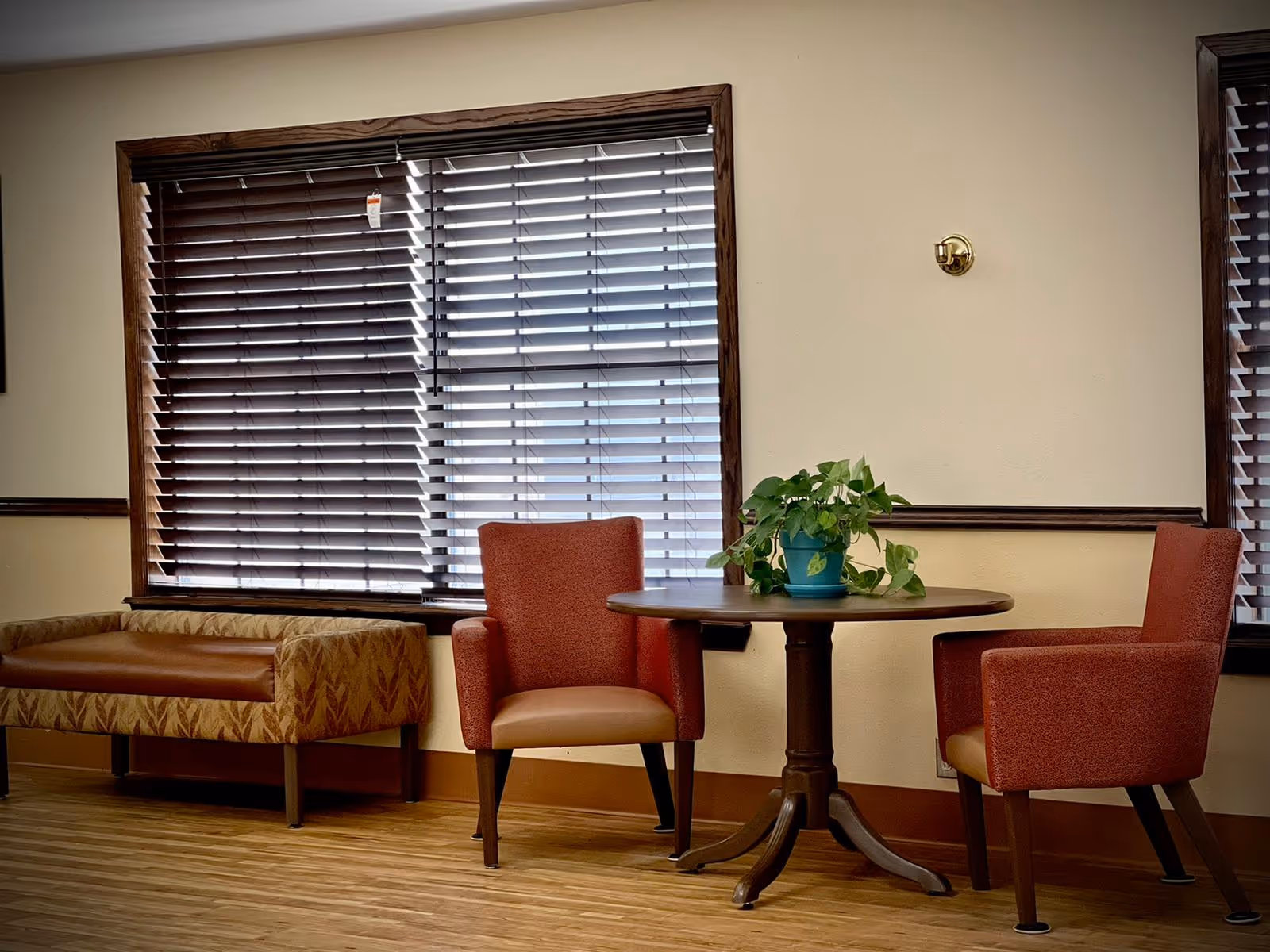 A cozy seating area with two red upholstered armchairs and a round wooden table between them, which holds a green potted plant. To the left, there is a cushioned bench with patterned upholstery. The background features two large windows with wooden blinds partially closed, and the floor is wood.