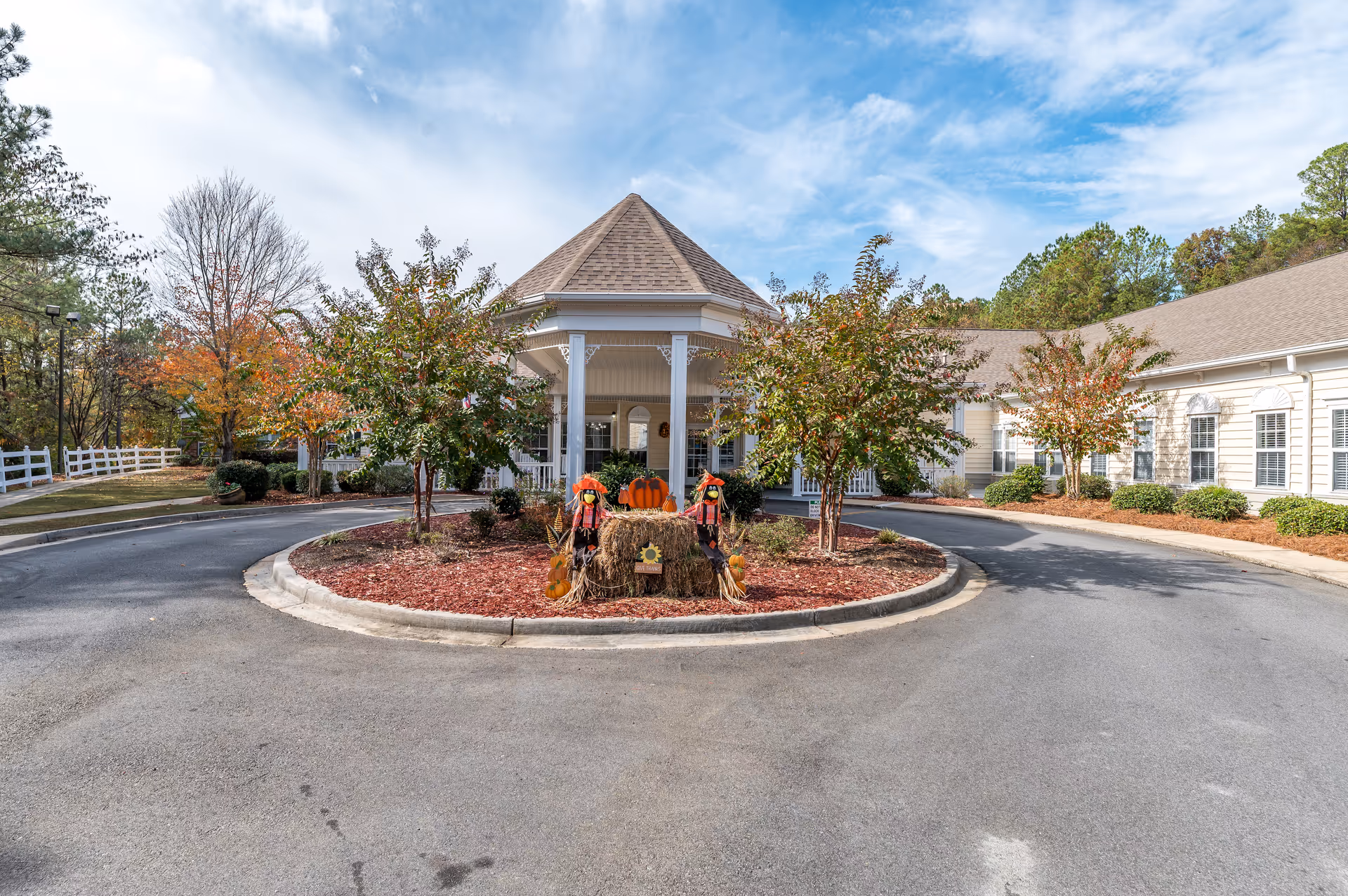 Entrance driveway of Oaks at Maple Ridge senior living facility featuring a circular landscaped island with small trees and fall-themed decorations including pumpkins and scarecrows. The building has a covered porch with white columns and beige siding, surrounded by trees with autumn foliage under a partly cloudy sky.