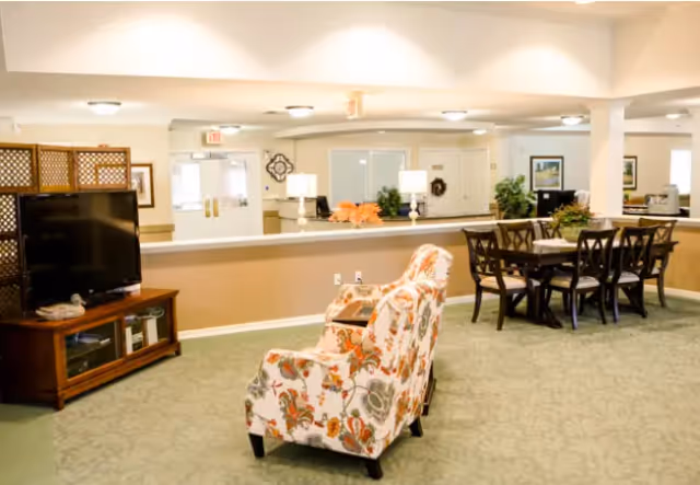 A spacious senior living common area with floral patterned armchairs, a wooden TV stand with a flat-screen television, a dining table with six chairs, and a reception desk in the background. The room is well-lit with ceiling lights and decorated with plants and framed pictures.