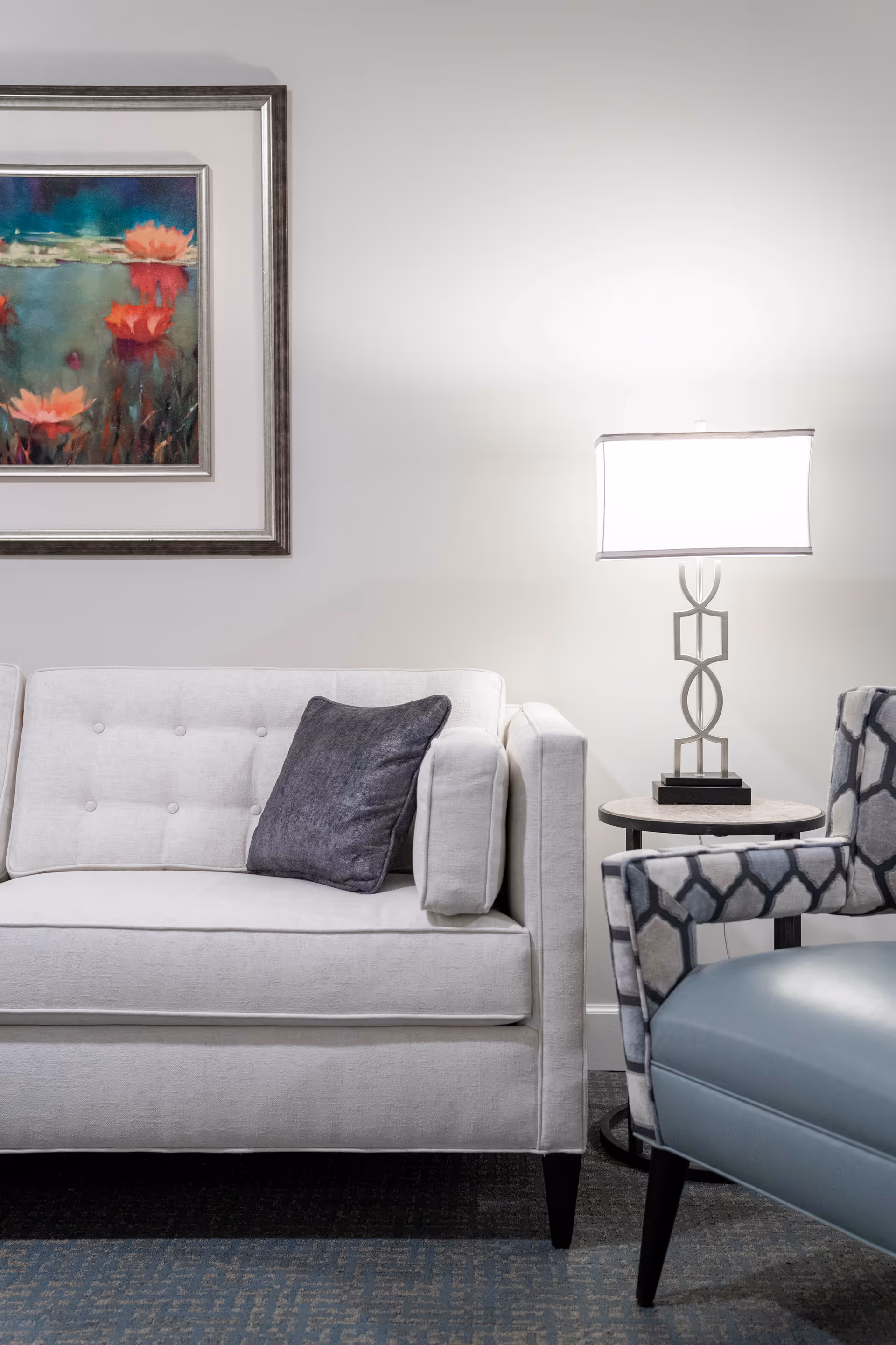A modern living room area with a white tufted sofa featuring a dark gray pillow, a patterned armchair with blue upholstery, a small round side table with a decorative lamp, and a framed painting of red and pink flowers on a greenish-blue background hanging on the wall.