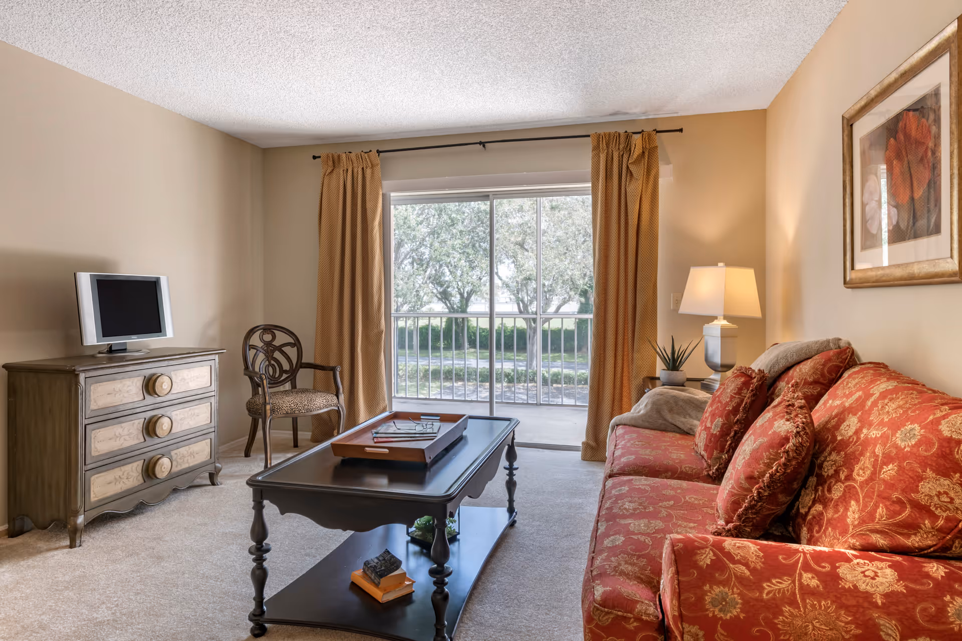 Living room with a red patterned sofa, coffee table, TV on a dresser, and sliding glass door leading to a balcony.