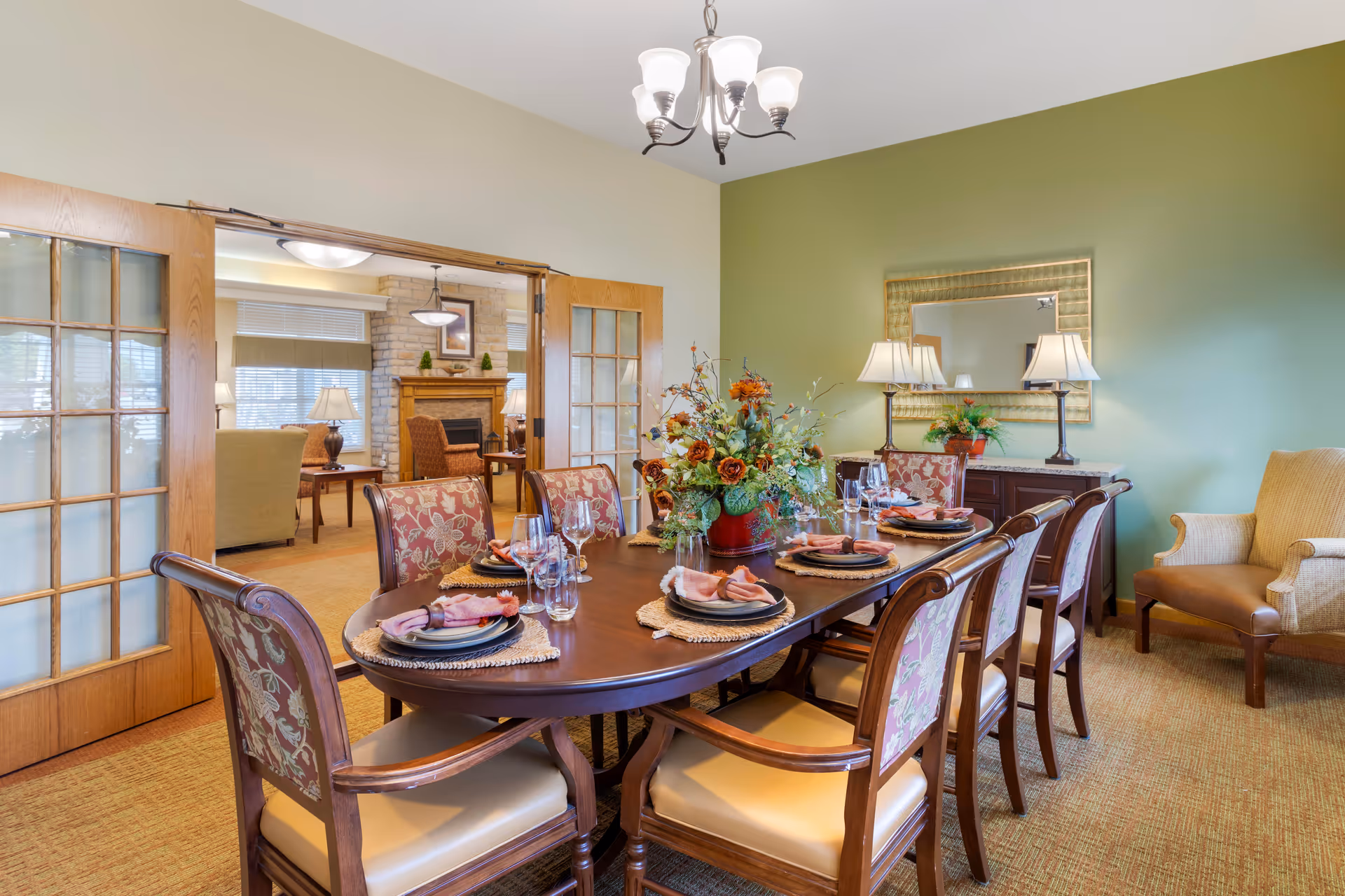 A dining room with a long wooden table set for six people with plates, napkins, glasses, and a floral centerpiece. The room features a green accent wall with a large mirror and two table lamps on a sideboard. There are upholstered chairs around the table and a beige armchair in the corner. French doors open to a living room area with a stone fireplace and additional seating.