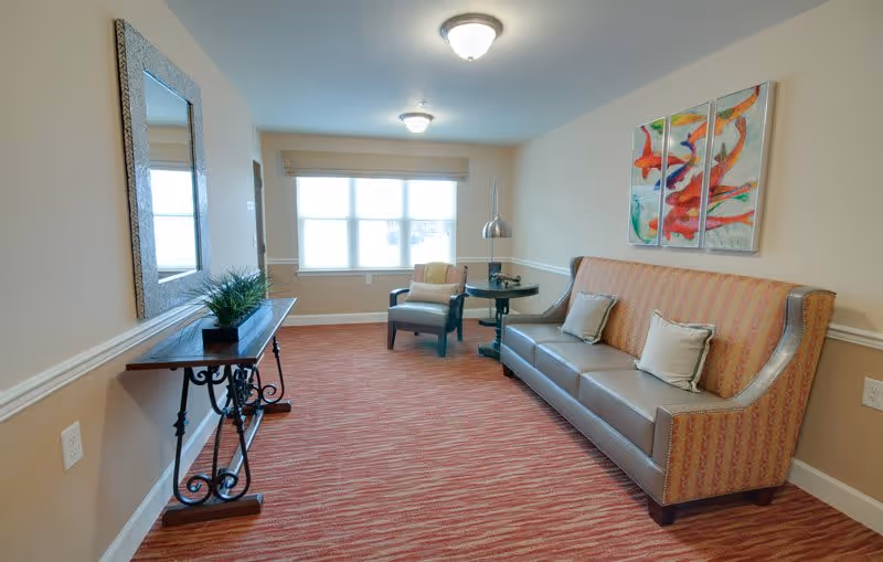 A well-lit sitting area in a senior living facility with a patterned sofa adorned with two cushions, a single armchair near a window, a small round side table with a modern lamp, a decorative console table with a potted plant, a large wall mirror, and colorful abstract artwork on the wall.