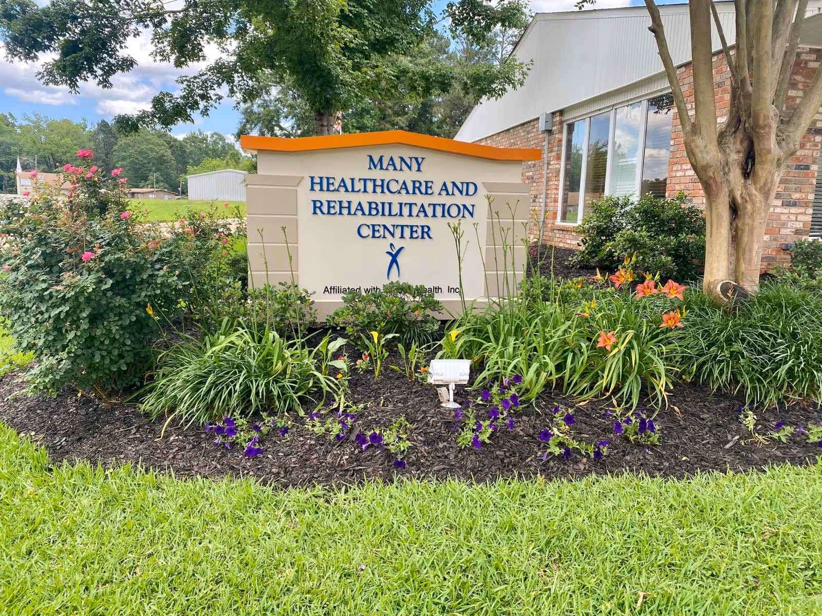 Flowerbed and landscaped lawn in front of a sign that reads 'Many Healthcare and Rehabilitation Center' by a brick building.