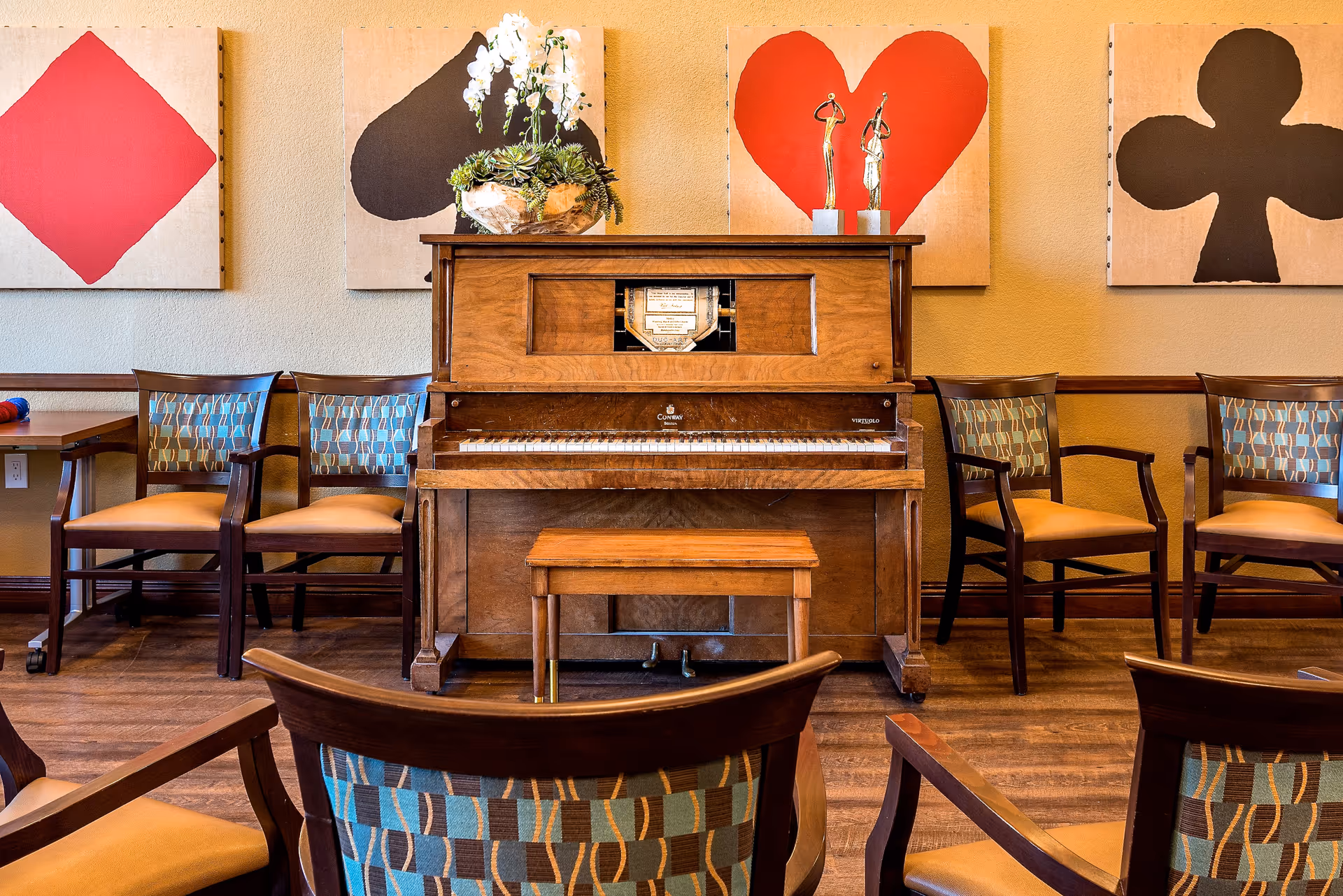 A cozy room with a wooden upright piano at the center, surrounded by several wooden chairs with patterned cushions. On top of the piano, there is a decorative plant and two small statues. The wall behind the piano features large paintings of playing card suits: diamond, spade, heart, and club.