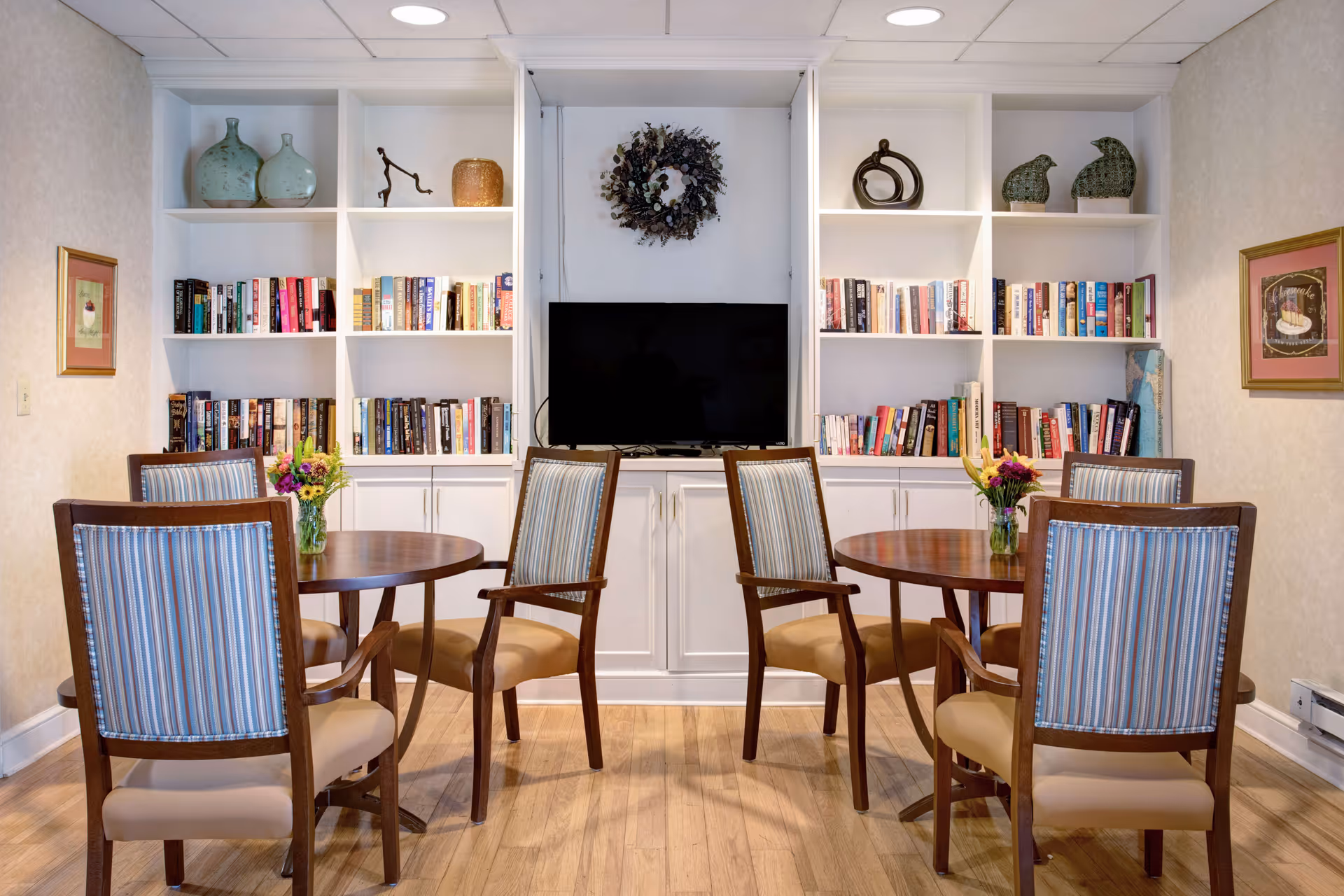 A cozy common area with two round wooden tables, each surrounded by four wooden chairs with striped upholstery. Behind the tables is a white built-in bookshelf filled with books and decorative items, with a flat-screen TV mounted in the center. The room has light-colored walls, wooden flooring, and framed artwork on either side of the bookshelf.