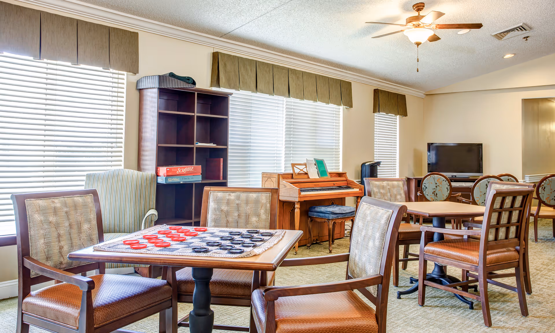 Bright communal activity room with tables and chairs, a checkers board, piano, shelving and a TV.