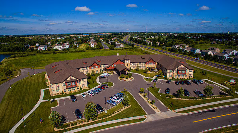 Aerial view of Guardian Angels Engel Haus Senior Living facility in Albertville, showing a large U-shaped building with a brown roof surrounded by parking lots, green lawns, and roads under a blue sky with scattered clouds.