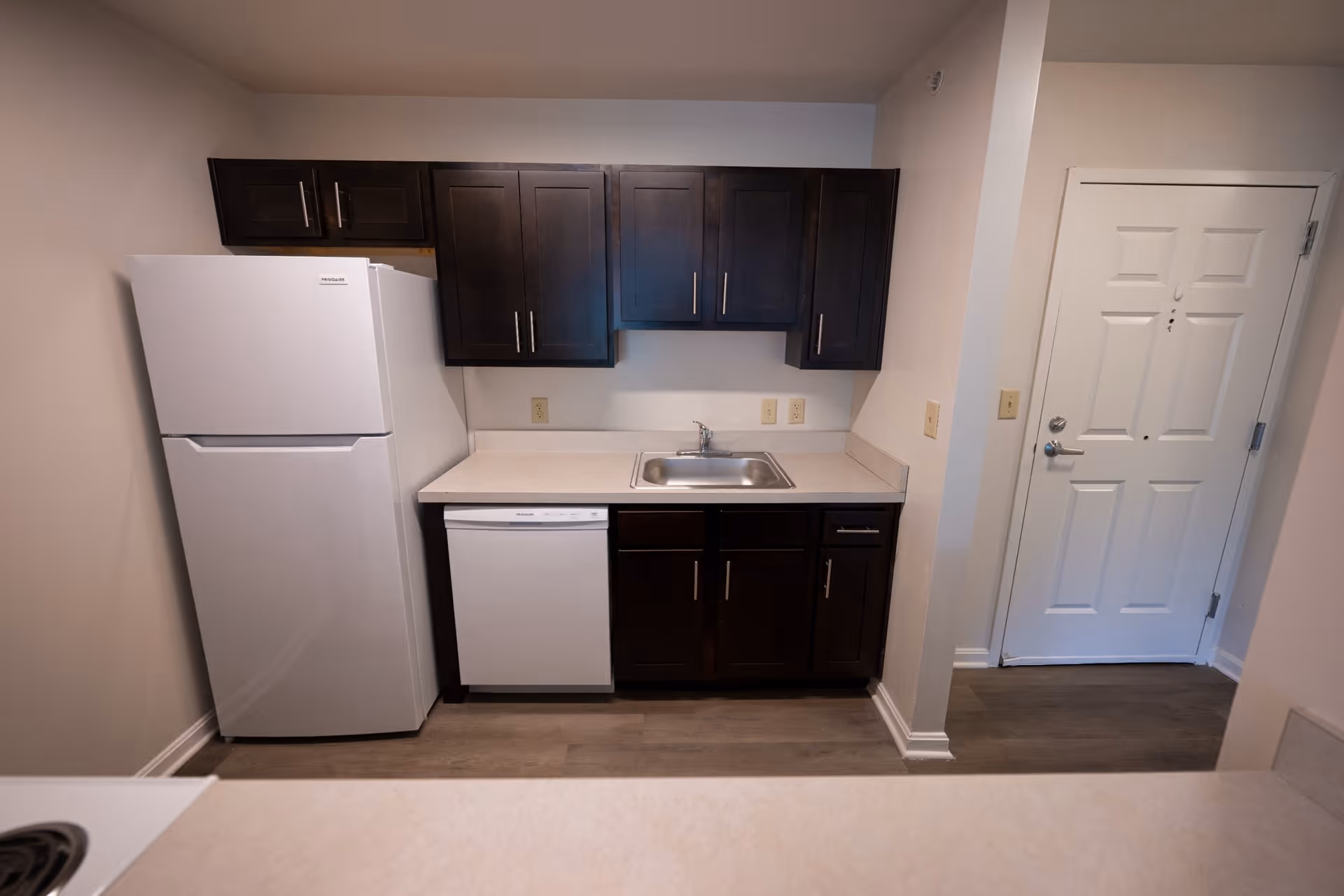 A small kitchen area featuring dark wood cabinets, a white refrigerator, a white dishwasher, a stainless steel sink, and a white countertop. To the right of the kitchen is a white door with a silver handle. The floor is a light wood laminate.