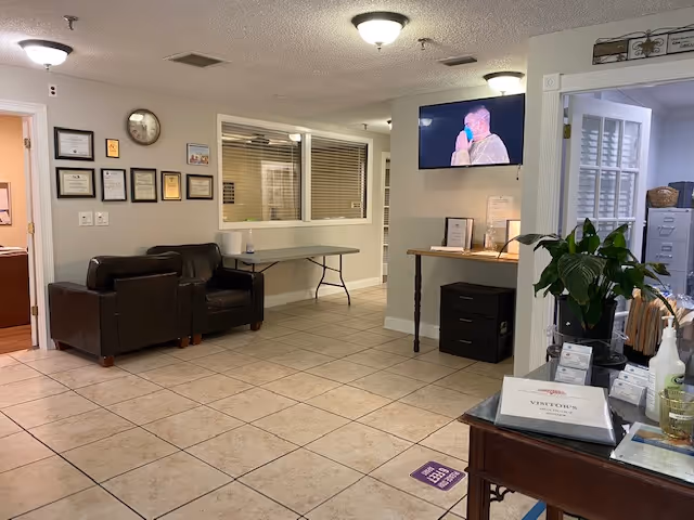 Interior reception area with leather chairs, tiled floor, a wall-mounted TV, and a check-in table with paperwork.