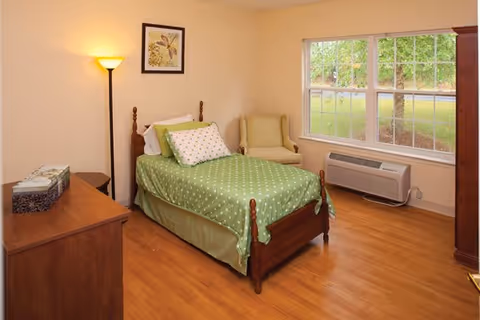 A tidy single bedroom with a wooden bed dressed in green polka-dot bedding, a striped armchair, dresser, floor lamp, and a large window overlooking greenery.
