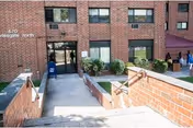 Exterior view of a brick building entrance with stairs and a ramp leading down to double glass doors. There are windows above the doors and some greenery near the entrance. A few people are visible on the right side near a purple canopy tent.