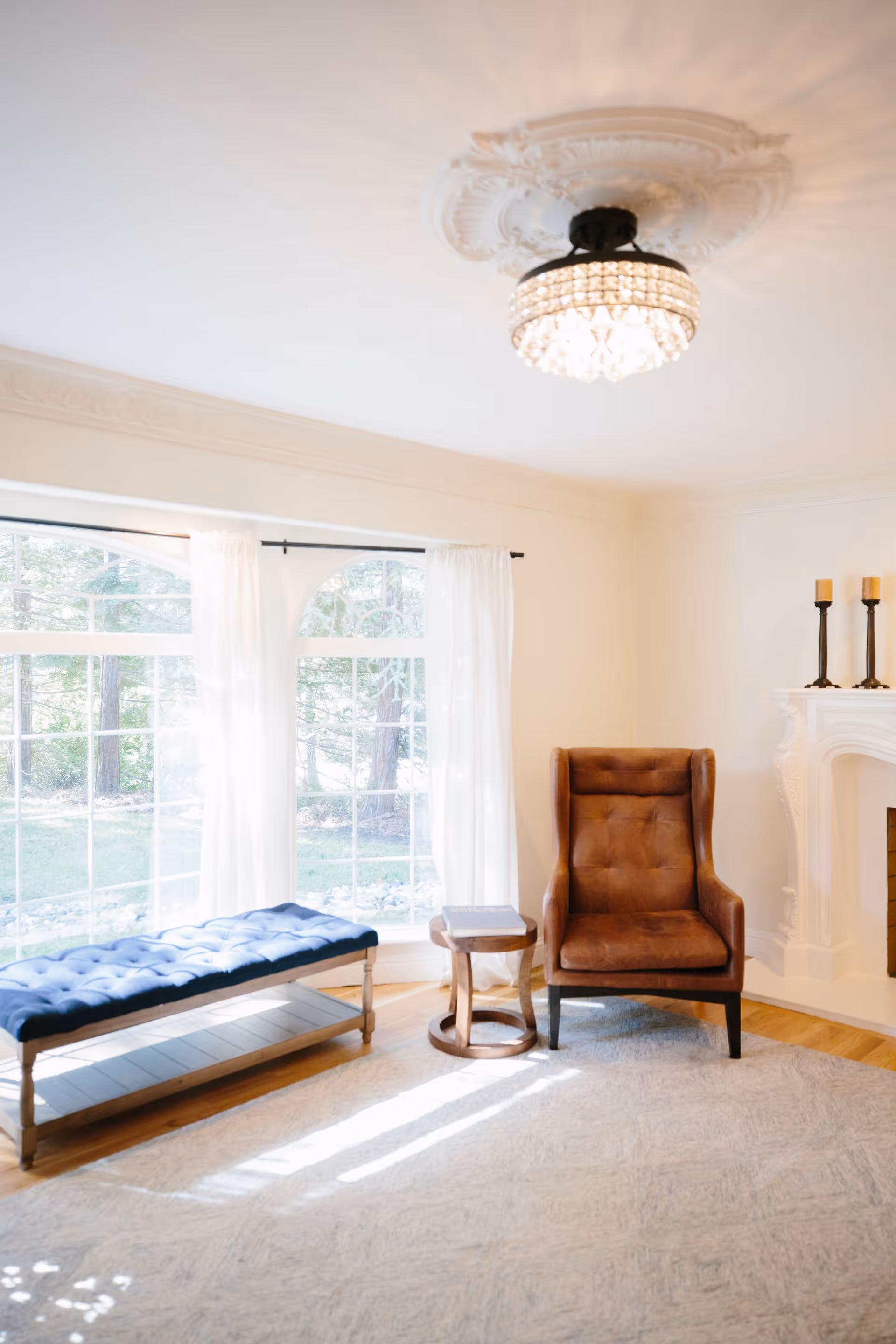 A bright living room with large windows covered by sheer white curtains, a tufted blue bench, a brown leather armchair, a small wooden side table with a book on it, a white fireplace with two candle holders, and a decorative ceiling light fixture.