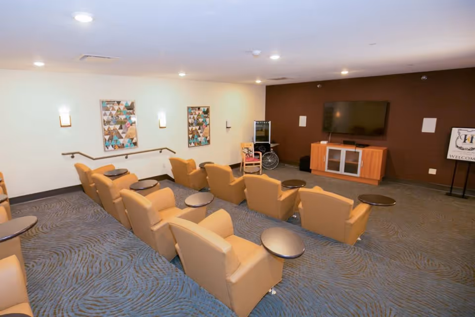 A cozy media room with two rows of tan recliner chairs, each equipped with a small round side table. The room has a blue patterned carpet, white and brown walls, two framed artworks, a large flat-screen TV mounted on the brown wall, and a wooden TV stand. There is also a wheelchair and a welcome sign near the TV stand.